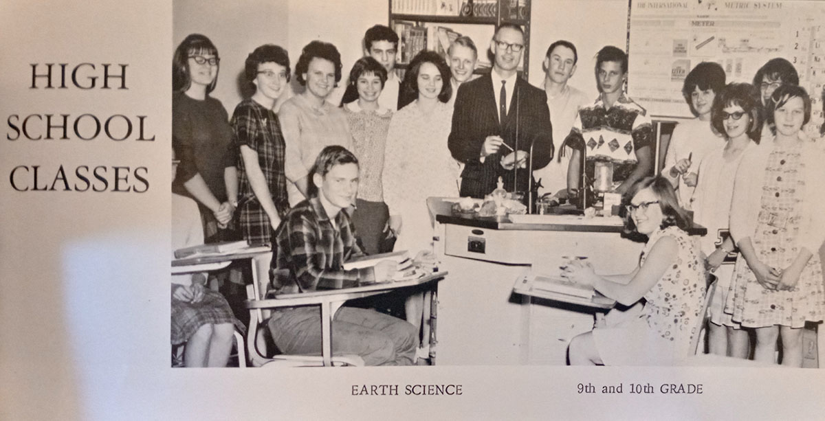 A yearbook photo of Mack as an Earth Science teacher.  Mack is standing in the center of the image in a dark suit, surrounded by sixteen high school students.  Some are sitting and others are standing.  THey are in a science classroom and Mack is standing behind a demonstration table with a sink.  The left of the image reads 'High School Classes' and the bottom of the image says 'Earth Science, 9th and 10th grade'.