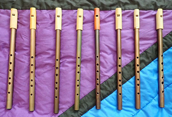 A view of eight copper whistles laid out vertically on a purple, blue, and black quilt geometric quilt background.  The whistle bodies are varying shades of brown.  Most have bamboo heads.